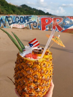 A photo of a pinapple filled with piña colada and topped with fruits, a little umbrella, and a small Puerto Rican flag. The pinapple is held in front of a beach mural that reads: Welcome to PR