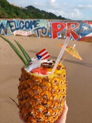 A photo of a pinapple filled with piña colada and topped with fruits, a little umbrella, and a small Puerto Rican flag. The pinapple is held in front of a beach mural that reads: Welcome to PR