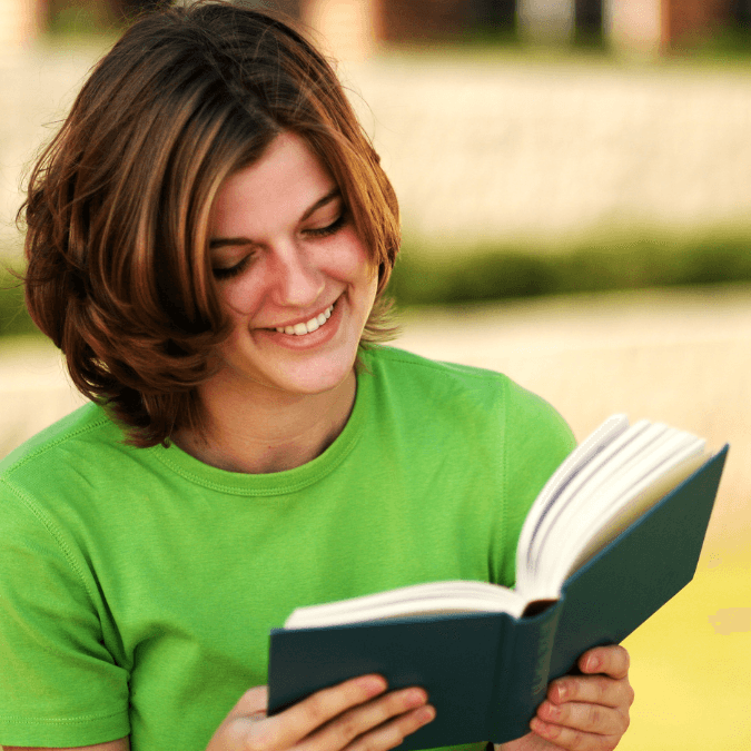 A woman with shoulder-length hair smiles while reading a book outdoors in a green shirt.